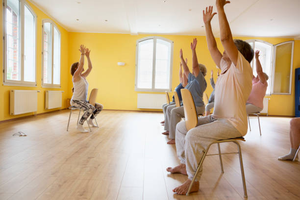 Teacher and active senior women yoga class on chairs Teacher and active senior women yoga class on chairs, arms raised, chair stock pictures, royalty-free photos & images