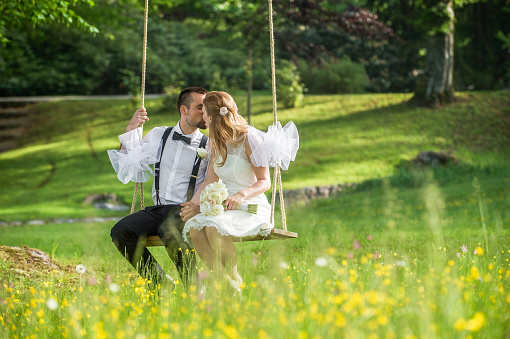 Bride and groom kissing each other while sitting on swing in park.