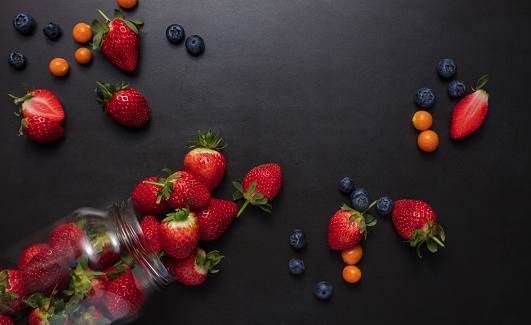 Fresh strawberries and blueberries on black background. Strawberries falling out of a glass jar.