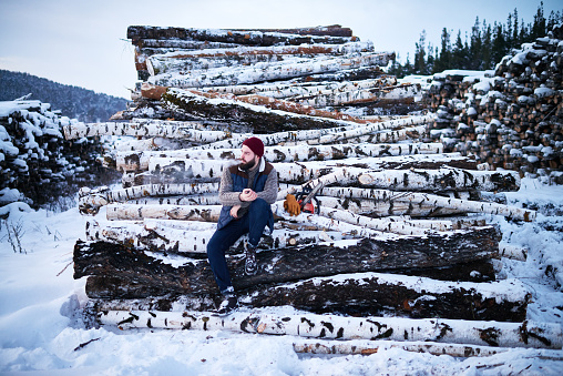 Pensive stylish young lumberjack in warm clothing smoking and contemplating while resting at workplace