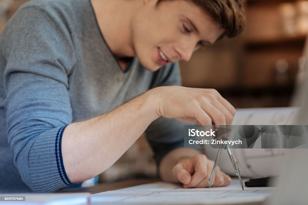 Charming man measuring distance with a pair of compasses Precise figures. The focus being on the hand of a handsome smiling young engineer holding a compass measuring a distance on a blueprint with it Activist Stock Photo