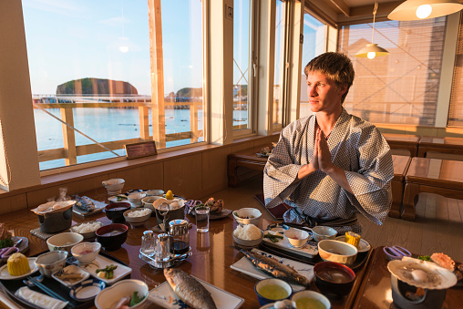 Young men in kimono sitting at the breakfast table with traditional Japanese food at sun rise. Lake in front of the window. Utoro, Japan.