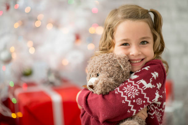 Hugging Teddy A young Caucasian girl is indoors in a living room on Christmas day. She is wearing festive clothing. She is sitting in front of a Christmas tree and hugging her new teddy bear. teddy bear photos stock pictures, royalty-free photos & images