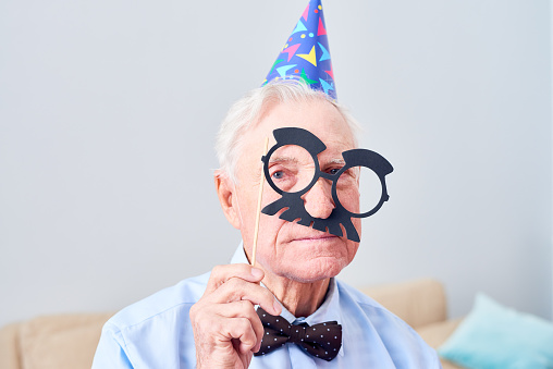 Unsmiling gray-haired senior man behind mask with glasses and mustache wearing party hat posing for camera