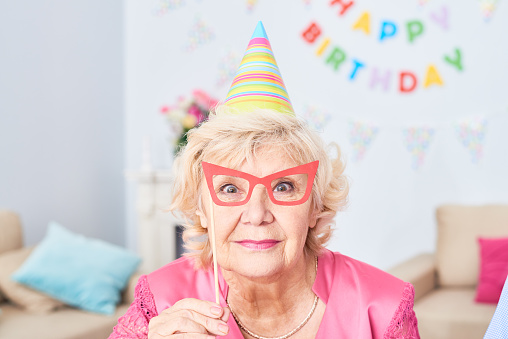 Smiling beautiful senior woman holding mask in shape of glasses on stick near face and looking at camera while spending time at birthday party at home