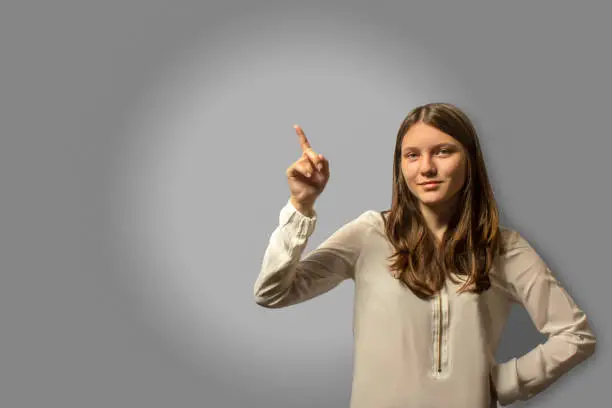 Photo of Portrait of a young beautiful student girl is attracted by attention with her indexed finger up. a lot of copy space. on a gray gradient background.