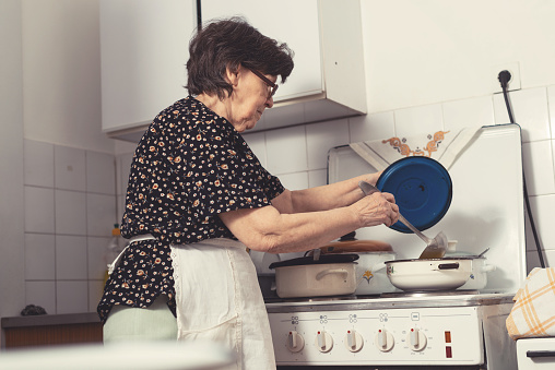 Cheerful senior woman from Eastern Europe cooking lunch for her family in old-fashioned kitchen. Portrait of active senior woman enjoying cooking.
