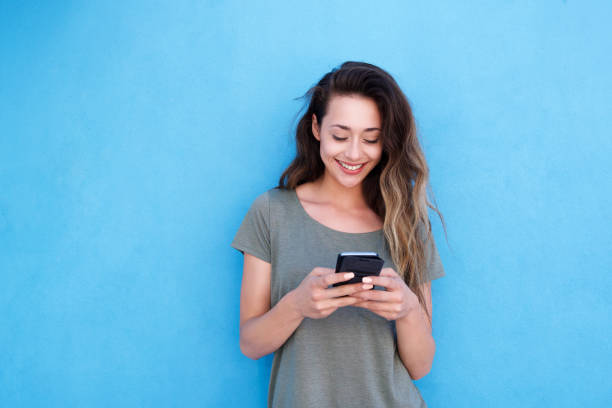 jovem mulher sorridente usando telefone celular contra o fundo azul - fashion one person relaxation cool - fotografias e filmes do acervo