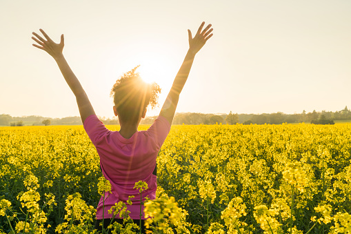 Mixed race African American girl female young woman athlete runner teenager in golden sunset or sunrise arms raised celebrating in field of yellow flowers