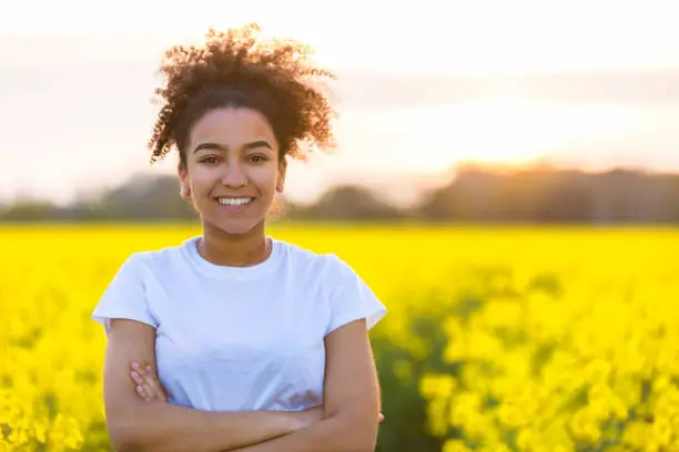 Photo of Beautiful mixed race African American girl teenager female young woman happy smiling with perfect teeth at the end of a path in a field of yellow flowers
