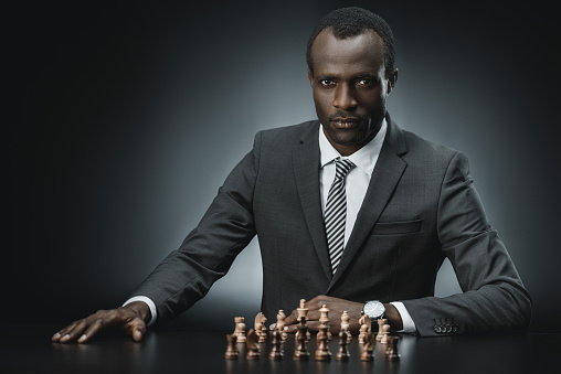 portrait of confident african american businessman with chess figures on table looking at camera isolated on black