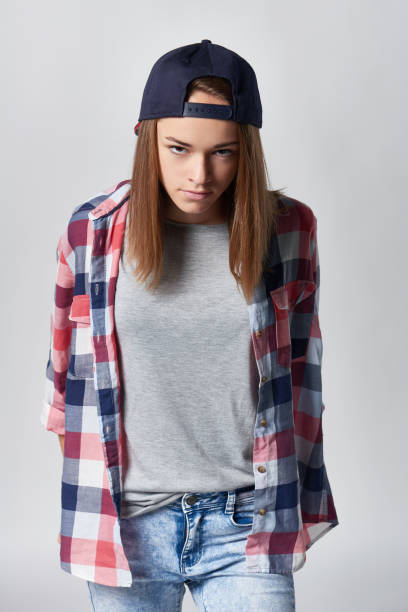 Angry teen girl wearing checkered shirt and baseball cap over grey background looking at camera sullen, front view