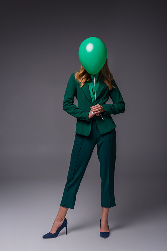 stylish girl in green suit holding green balloon in front of face, on grey