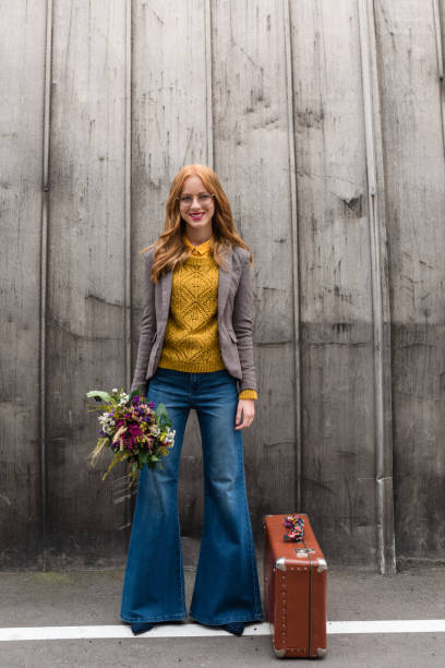 smiling girl with flowers and suitcase attractive smiling stylish redhead girl with bouquet of flowers and vintage suitcase flare pants stock pictures, royalty-free photos & images