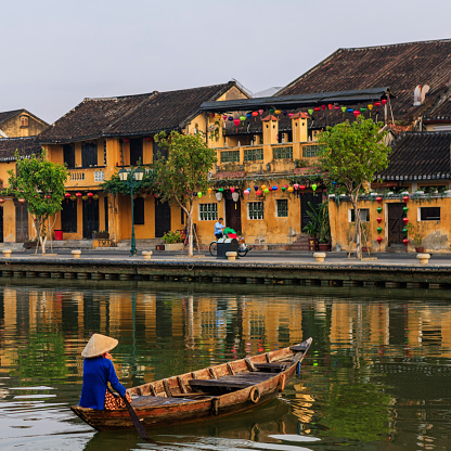 Vietnamese woman paddling in old town in Hoi An city, Vietnam. Hoi An is situated on the east coast of Vietnam. Its old town is a UNESCO World Heritage Site because of its historical buildings.