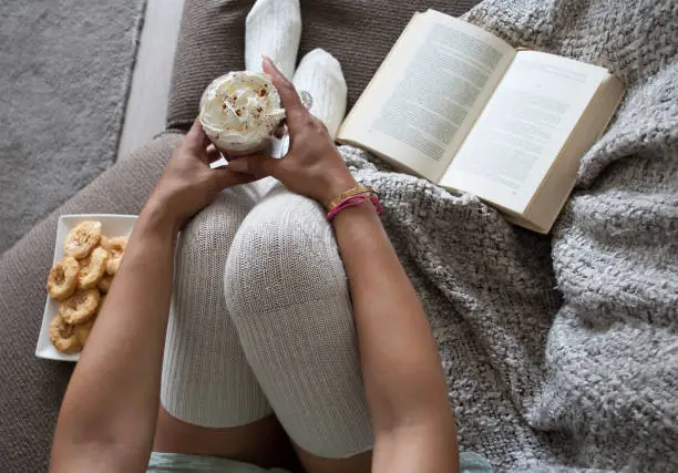 closeup of woman reading book at home on couch with hot chocolatemilk and cookies, top view