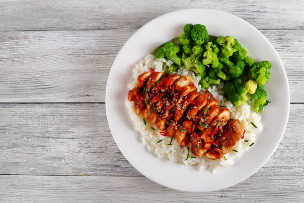 white rice topped with teriyaki chicken white rice topped with teriyaki chicken breast served with steamed broccoli on white plate. teriyaki sauce in gravy boat, chili peppers and parsley on wooden worktop, view from above sticky sesame chicken sauces stock pictures, royalty-free photos & images