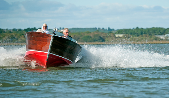A stylish retro couple turning their boat at speed, in a pristine classic gentlemans highly polished wooden powerboat, reminiscent of the art deco nineteen twenties style of a bygone era, Poole Harbour, England, UK