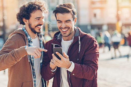 Two young men looking at a smart phone and laughing outdoors in the city