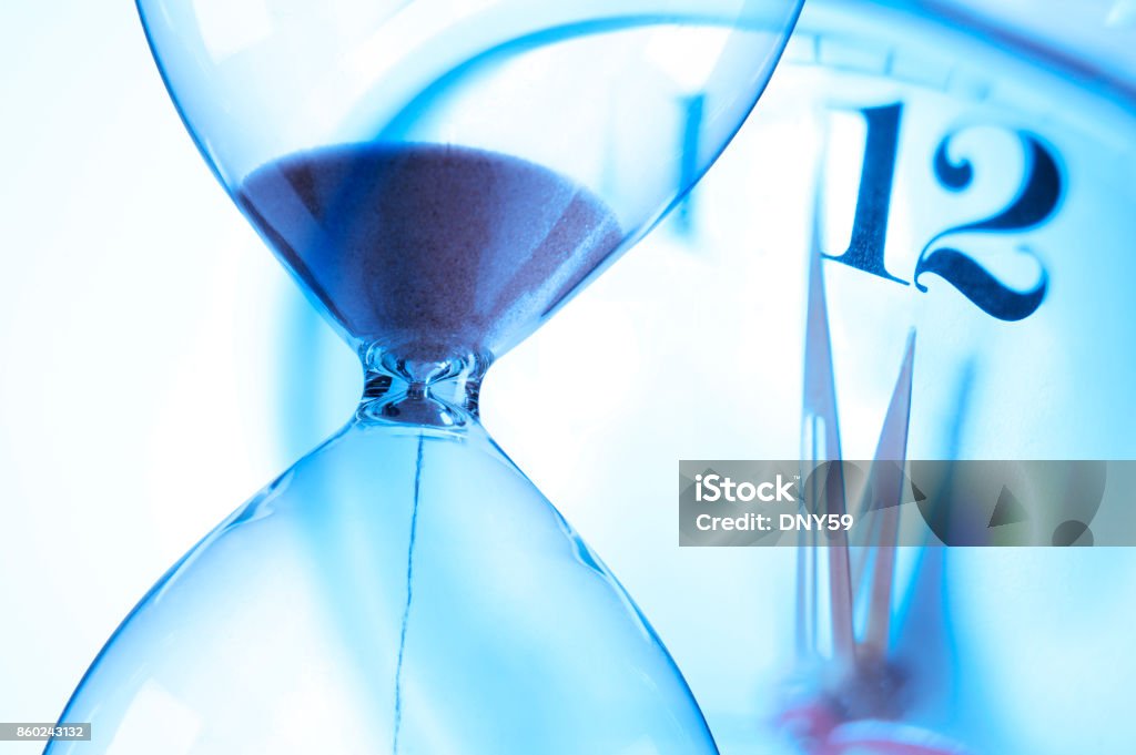 Hourglass And Clock About To Strike Twelve O'Clock Sand pours through an hourglass as a clock in the background is about to strike 12 o'clock. Deadline Stock Photo