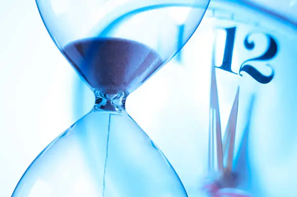 Sand pours through an hourglass as a clock in the background is about to strike 12 o'clock.