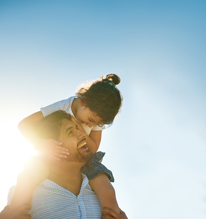 A young mixed race girl sits on her dad's shoulders, looking at each other, both smiling outdoors in the sun