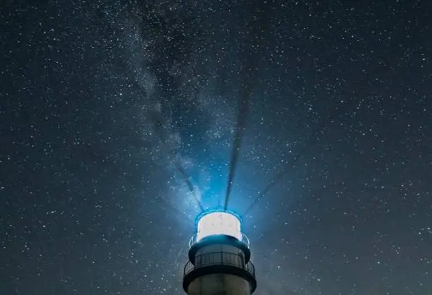 Photo of Lighthouse at night with radiating beams and starry sky with Milky Way