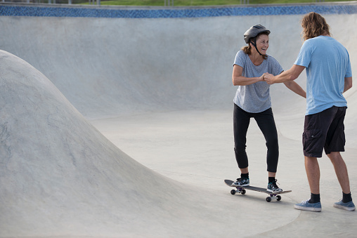 Skateboard coaching and skills training. Active senior woman taking a skateboard lesson, trying to keep balance with trainer/coach support.