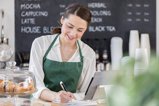 Cheerful Hispanic female barista writes an order while working behind the counter at a trendy coffee shop.