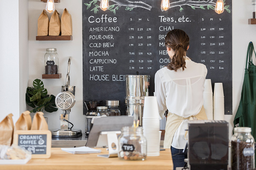 Rear view of female barista getting coffee for a customer in a trendy coffee shop. She is standing in front of a chalkboard men.