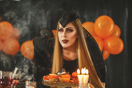 Young woman dressed as vampire is standing by the table decorated in Halloween spirit.