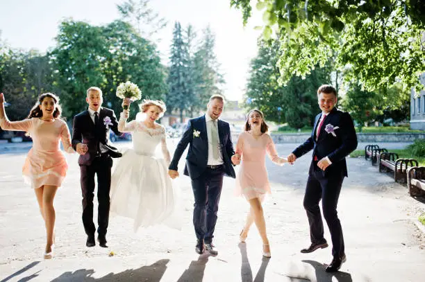 Happy wedding couple and bridesmids with groomsmen running and jumping on a sunny day in the park.