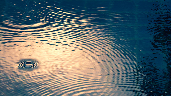 Rain drop falling on swiming pool and ripple shape and sun light reflection on water surface.