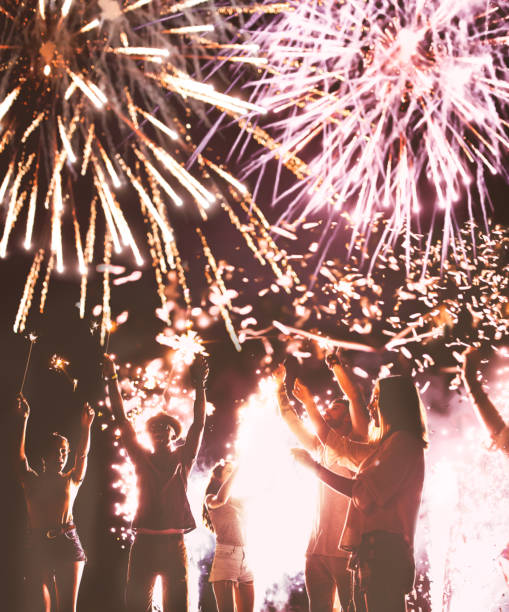 Young multi-ethnic crowd celebrating Fourth of July with sparklers and fireworks display in night sky