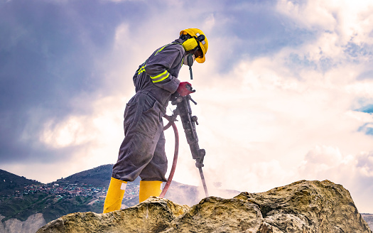 Worker with helmet and protective suit using a drilling machine on top of a large rock