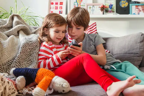 Children in living room with sitting on a sofa. The older brother is showing the little girl his mobile phone.