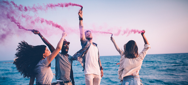 Young multi-ethnic hipster friends on summer holidays celebrating with smoke bombs on the beach