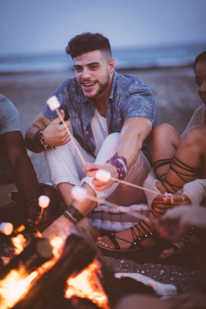 istock Young hipster man roasting marshmallows over bonfire at beach party 860595270