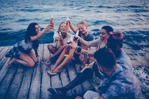 Happy multi-ethnic hipster friends celebrating, drinking beer, toasting with beer bottles and sitting on jetty