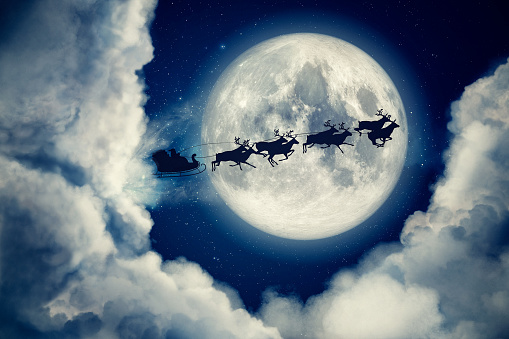 Blue xmas eve night with moon and clouds with Santa Claus sleight and reindeer silhouette flying to bring gifts and presents with text space to place logo or copy. Christmas present greeting post card.