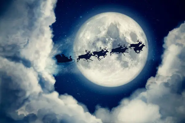 Photo of Blue xmas eve night with moon and clouds with Santa Claus sleight and reindeer silhouette flying to bring gifts and presents with text space to place logo or copy. Christmas present greeting post card