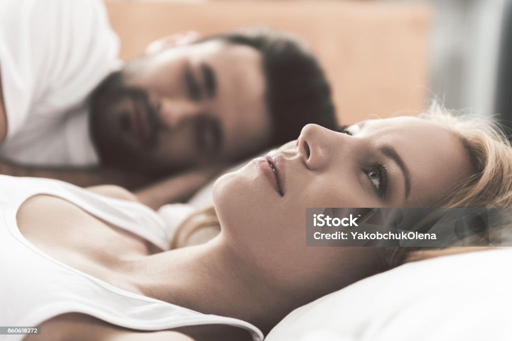 Girl worries about her marriage Nervous young woman is lying on bed and thinking seriously. Her husband is sleeping on background Couple - Relationship Stock Photo