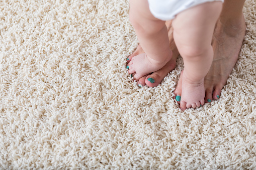 Unrecognizable baby walks on her mom's feet on the carpet. Only their feet are shown in the photo.