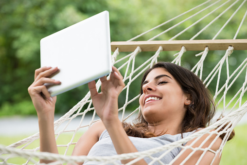 Smiling Young Woman Lying On Hammock Looking At Digital Tablet