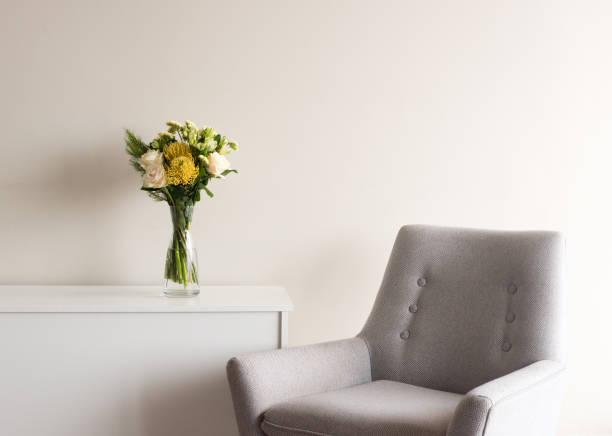 Close up of grey retro armchair next to white cabinet with cream and yellow flowers in glass vase against neutral wall background