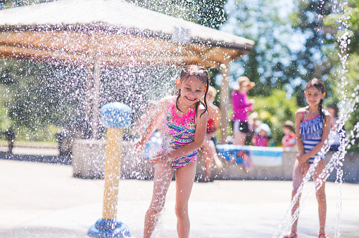 A group of young children frolic and run in a playground splash pad. It is a sunny, perfect day for getting wet and playing hard! A young girl in a swimsuit is running through the sprinklers and yelling and laughing as the water droplets cascade all around and her friends wait in the background.