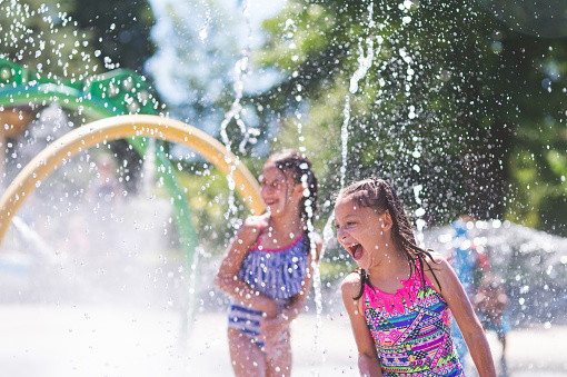 A group of young children frolic and run in a playground splash pad. It is a sunny, perfect day for getting wet and playing hard! Two young girls in swimsuits are running through the sprinklers and yelling and laughing as the water droplets cascade all around them.