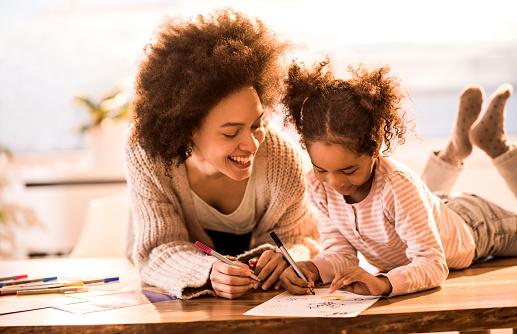 Young black mother assisting her little daughter in drawing at home.