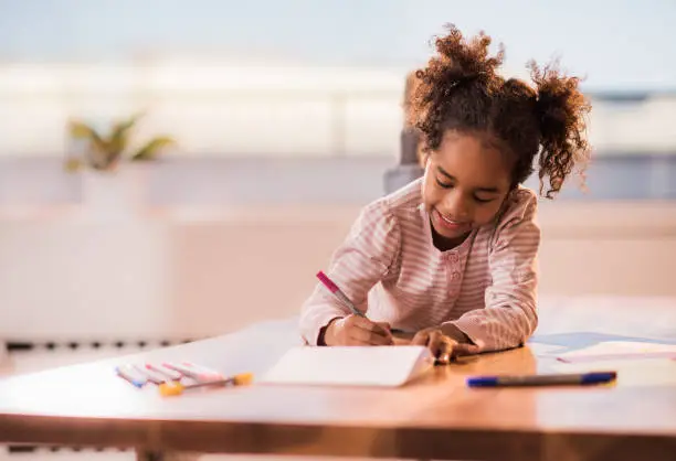 Happy African American girl relaxing on the table at home and coloring in notebook.