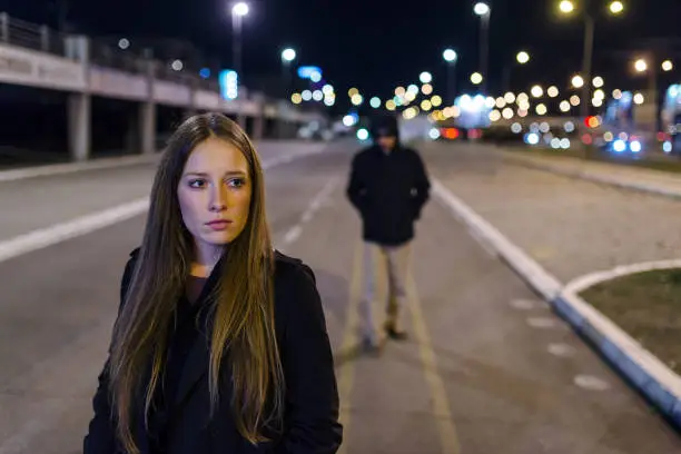 Beautiful young woman walking and being stalked by man criminal on the street at night. Dangerous situation for lonely female. Unrecognizable male figure with hidden face in hood walking, looking dangerous, stalking night robber burglar, bad troubled period, hooded guy Following frightened woman